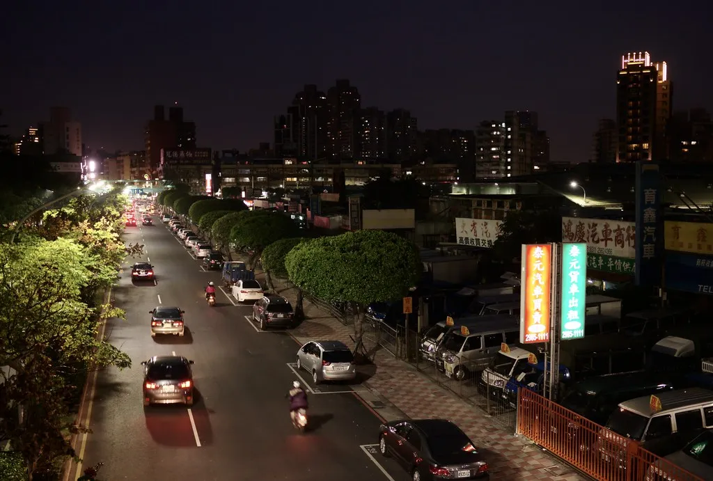 A tree-lined avenue in New Taipei City at night with cars, scooters and lit shop signs